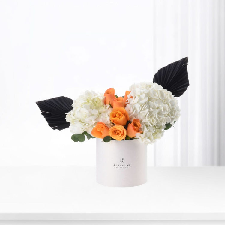 White cylindrical box holds vibrant orange roses and white hydrangeas, accented with dramatic black fan leaves.