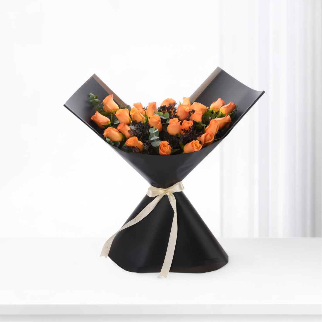 Vibrant orange roses in sleek black paper, accented with cream ribbon and dark foliage.