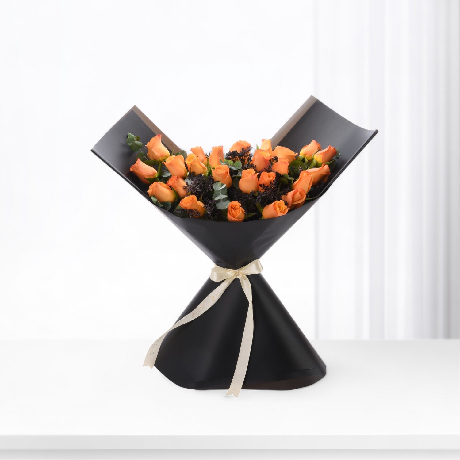 Vibrant orange roses in sleek black paper, accented with cream ribbon and dark foliage.