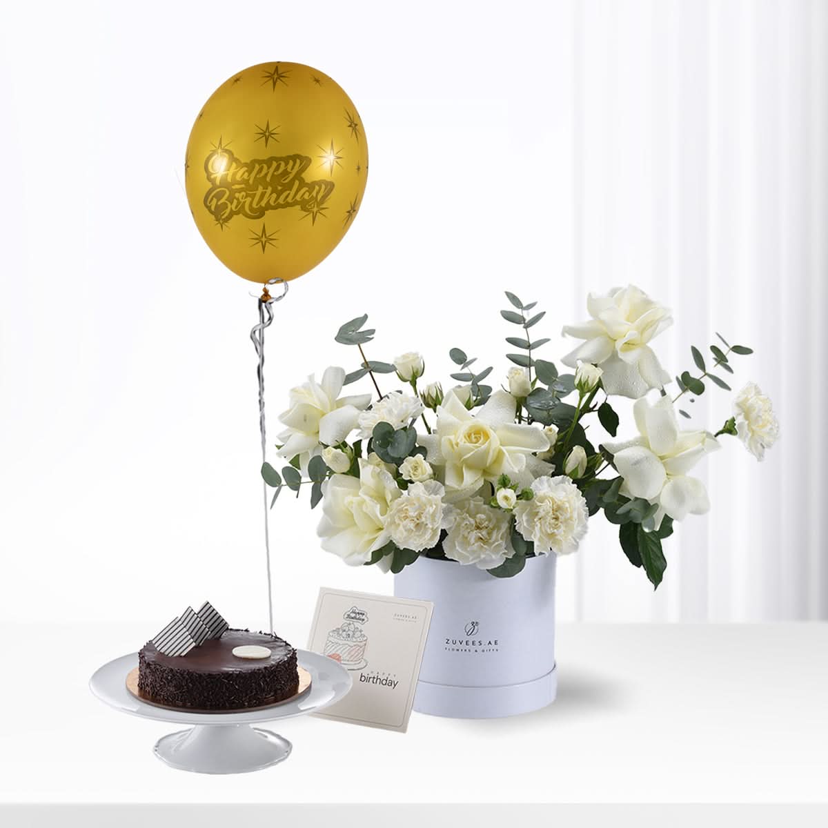 Golden "Happy Birthday" balloon floats beside white floral arrangement and chocolate cake.