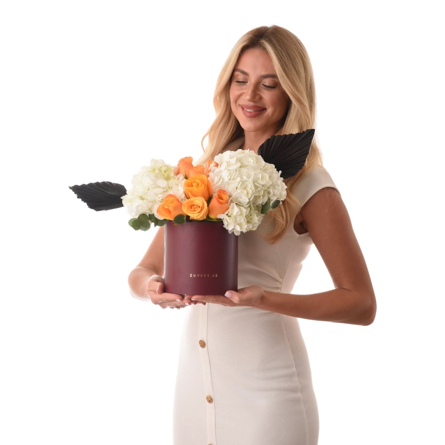 Woman holds burgundy box with orange roses and white hydrangeas, accented by black fan leaves.
