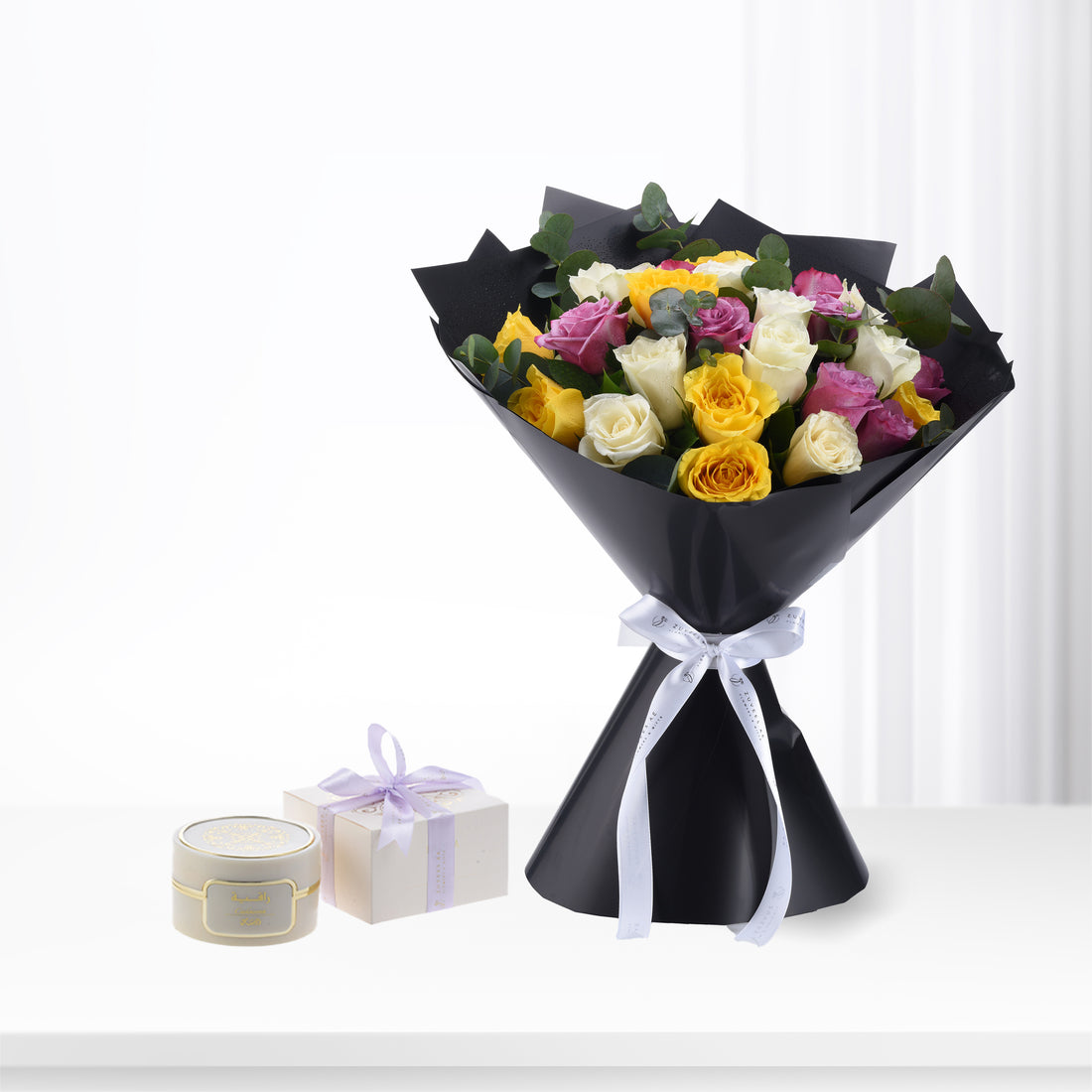 Vibrant yellow, pink, white roses in sleek black wrap with silver ribbon, beside gift boxes.