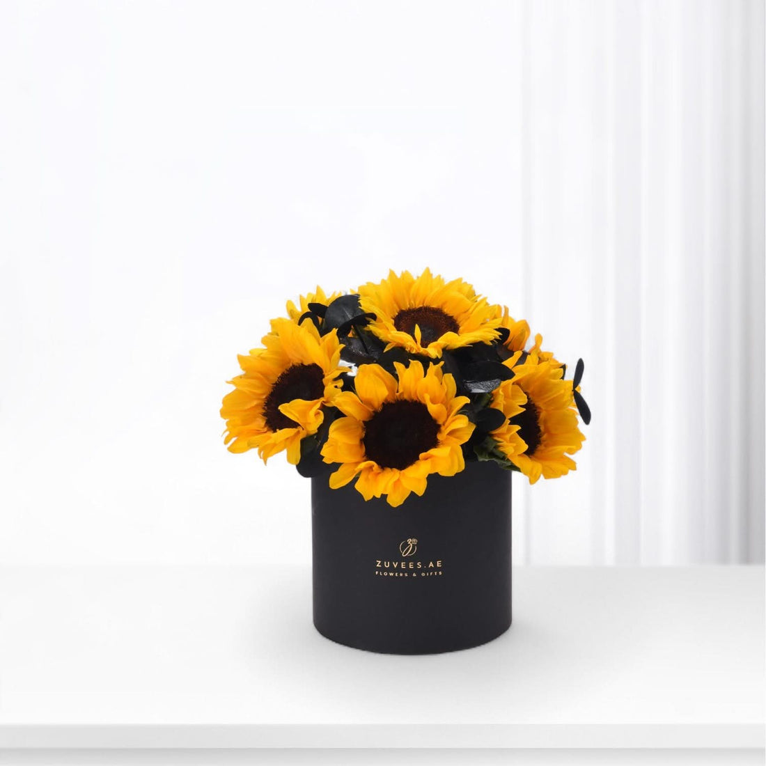Vibrant sunflowers in matte black cylindrical vase with gold logo.