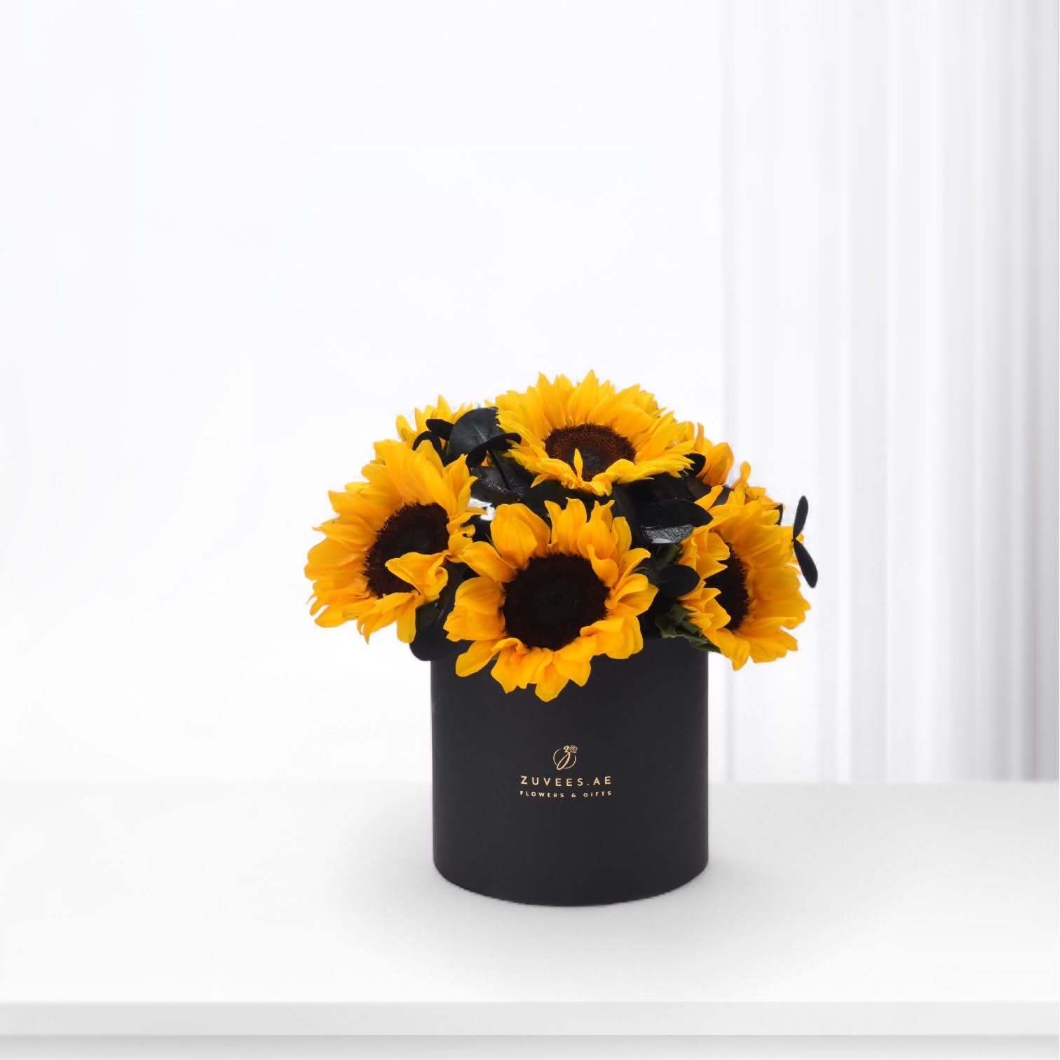 Vibrant sunflowers in matte black cylindrical vase with gold logo.