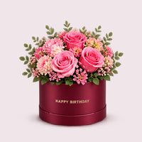 A luxurious cylindrical burgundy hatbox overflowing with a vibrant mixed bouquet of pink, coral, and magenta roses, complemented by delicate filler flowers and green eucalyptus foliage. The box features gold lettering reading 'HAPPY BIRTHDAY'