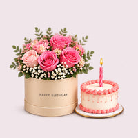 A cream-colored cylindrical hatbox with 'HAPPY BIRTHDAY' embossed text, filled with a vibrant mixed floral arrangement of hot pink and coral roses, pink carnations, white baby's breath, and green eucalyptus foliage. Beside it sits a classic layer cake with white frosting, pink piped rosette border details, and a single lit pink birthday candle on top.