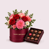 A burgundy cylindrical hatbox with 'HAPPY BIRTHDAY' in gold lettering, filled with a vibrant mixed floral arrangement of deep red roses, pink dahlias, coral carnations, white filler flowers, and green eucalyptus. Beside it sits an open burgundy chocolate box containing assorted truffles and chocolates in brown and dark varieties.