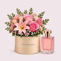 A cream-colored cylindrical hatbox with 'HAPPY BIRTHDAY' embossed in gold lettering, filled with a lush floral arrangement featuring pink roses, a pale pink lily, coral carnations, white baby's breath, and green eucalyptus foliage. Beside the arrangement stands a luxury perfume bottle with a rose-gold cap and pale pink fragrance.