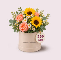 A cream-colored cylindrical hatbox with 'HAPPY BIRTHDAY' embossed text, filled with a cheerful mixed floral arrangement featuring two bright yellow sunflowers, coral pink roses, white daisy filler flowers, and green eucalyptus foliage. A circular price tag reading '299 AED' hangs from the box.