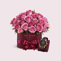 A burgundy cylindrical hatbox with 'HAPPY BIRTHDAY' in gold lettering and a matching burgundy silk ribbon bow, overflowing with a lush arrangement of mauve and pink roses with green foliage. Beside it sits a luxury chronograph watch with a black leather strap and rose-gold accents on a matching burgundy display block