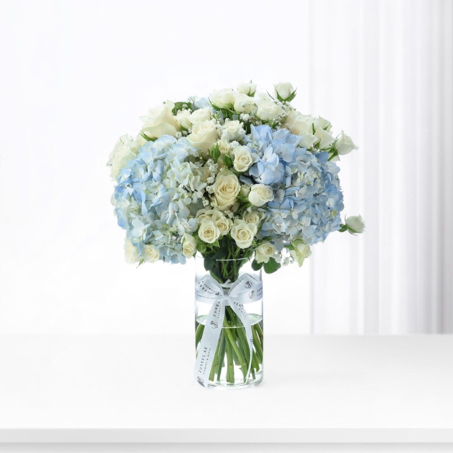 Elegant blue hydrangeas and white roses in clear vase with branded ribbon.