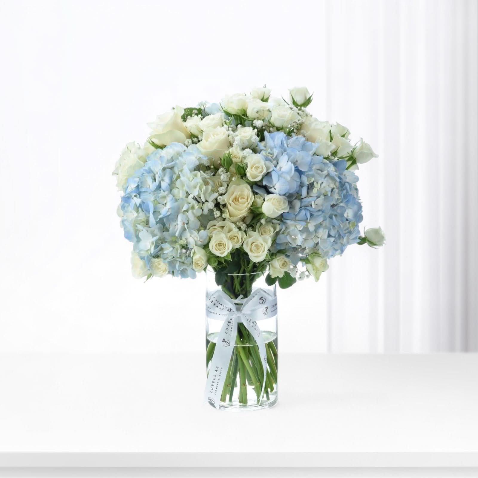 Elegant blue hydrangeas and white roses in clear vase with branded ribbon.