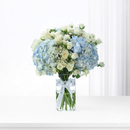 Elegant blue hydrangeas and white roses in clear vase with branded ribbon.