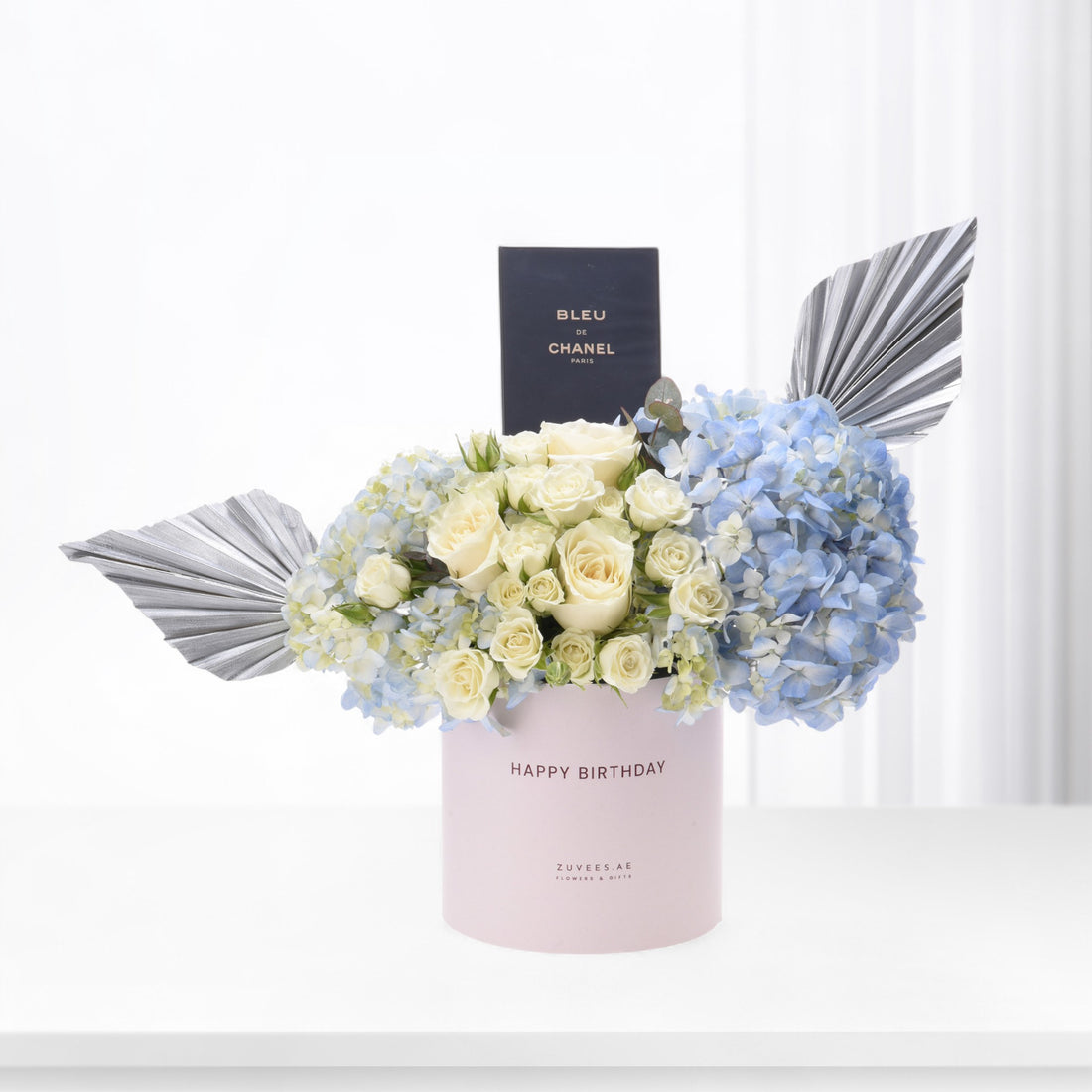 Pastel pink box holds creamy roses and blue hydrangeas, accented with silver fan leaves and a Chanel perfume box.
