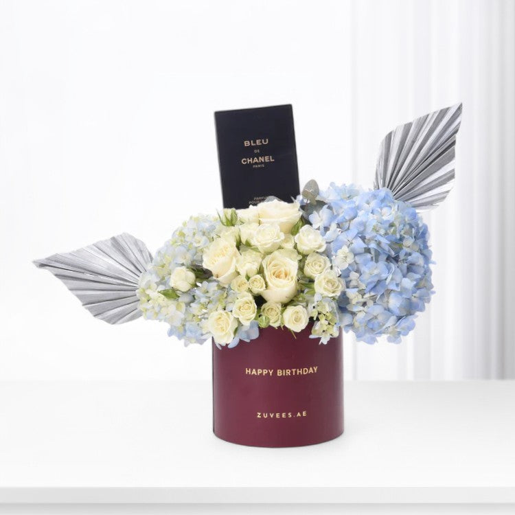 Maroon cylinder holds creamy roses, blue hydrangeas, and Chanel perfume—elegant birthday gift.