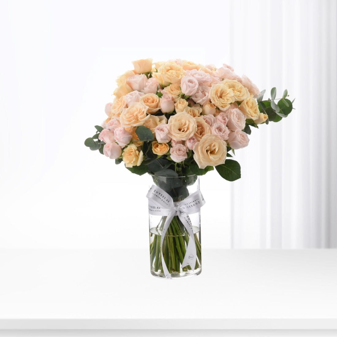 Pastel peach roses in clear vase, white ribbon bow, minimalist white background.