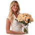 Woman holds lush peach rose bouquet in clear vase with branded ribbon.