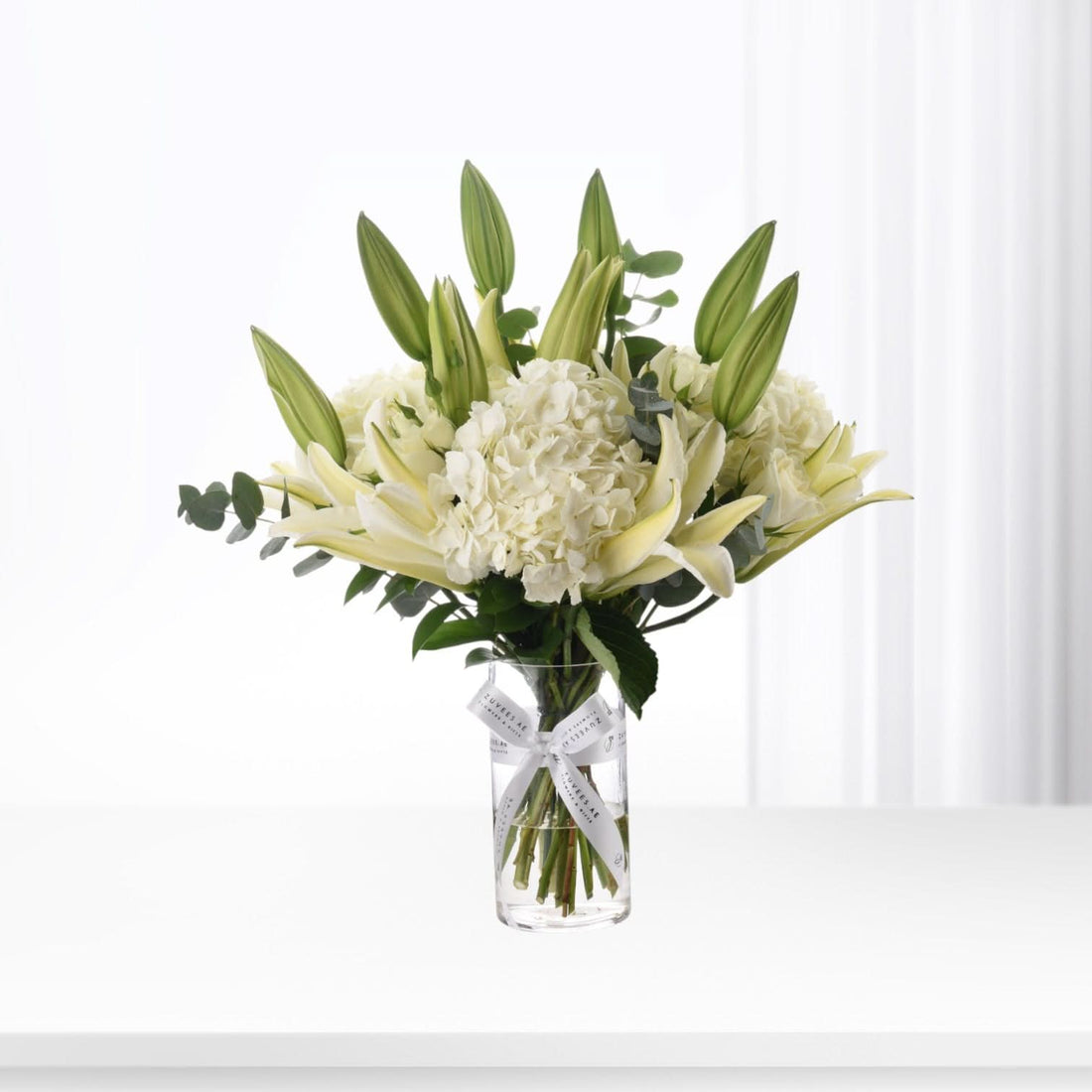 Elegant white lilies and hydrangeas in clear vase with branded ribbon.