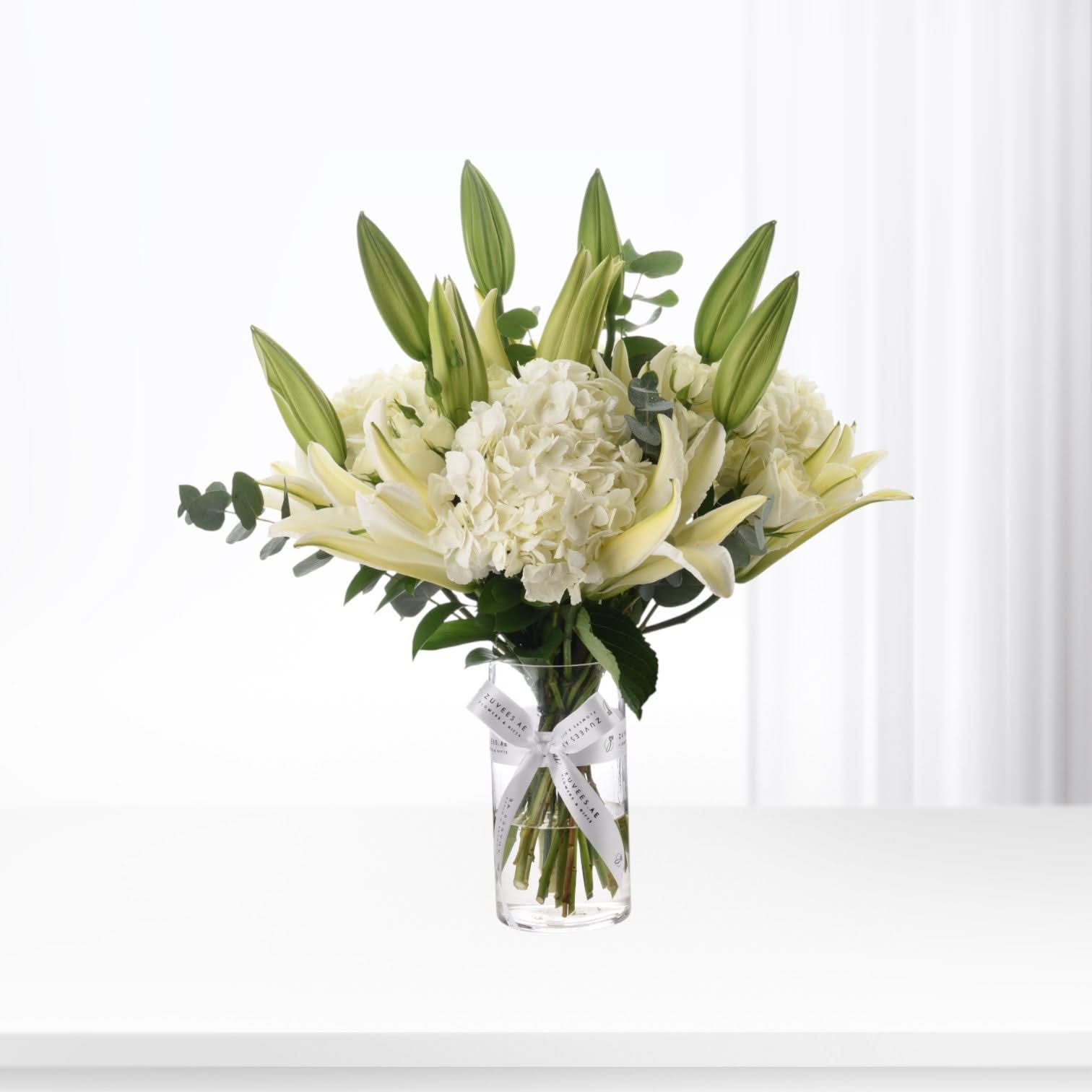 Elegant white lilies and hydrangeas in clear vase with branded ribbon.