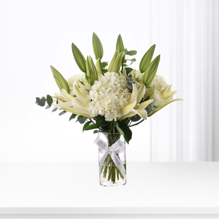 Elegant white lilies and hydrangeas in clear vase with branded ribbon.