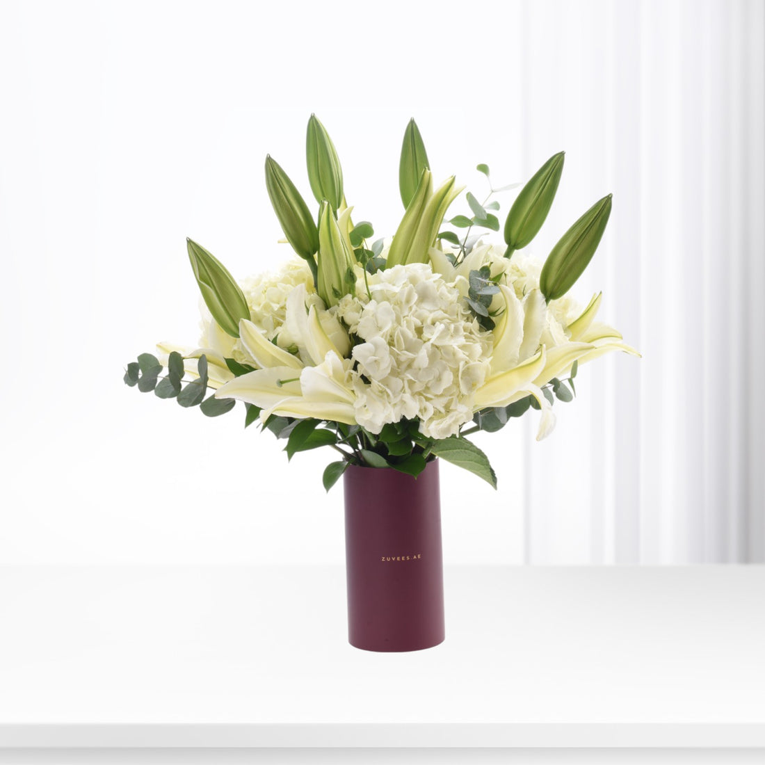 Elegant white lilies and hydrangeas in burgundy vase against soft white backdrop.