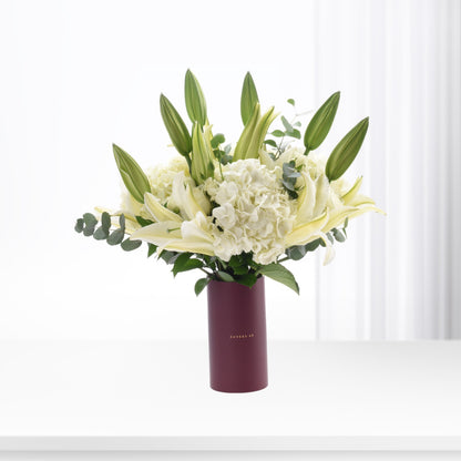 Elegant white lilies and hydrangeas in burgundy vase against soft white backdrop.