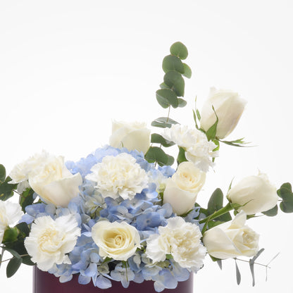 Elegant white roses, carnations, blue hydrangeas, and eucalyptus in a burgundy vase.