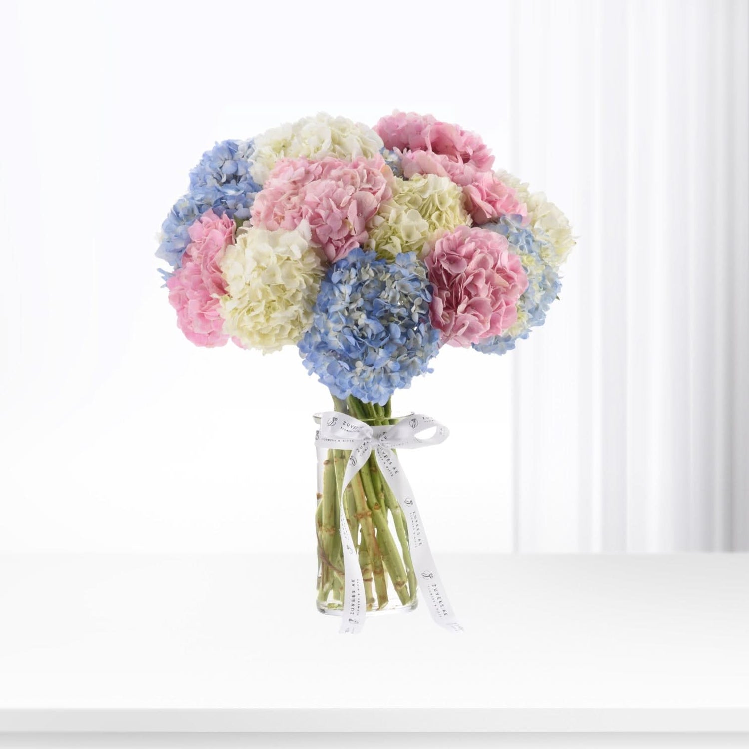 Pastel pink, blue, and cream hydrangeas in clear vase, tied with branded ribbon.