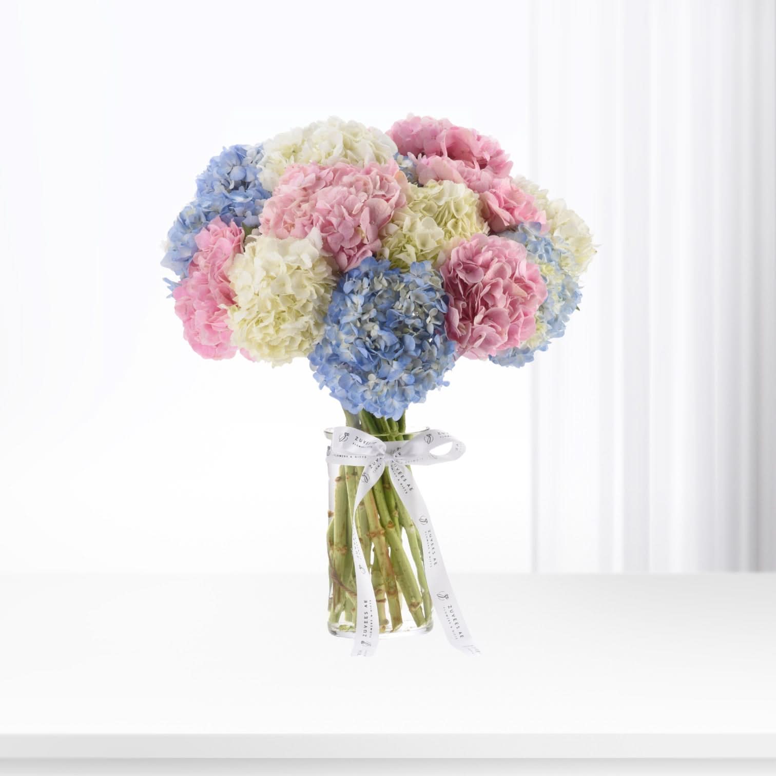 Pastel pink, blue, and cream hydrangeas in clear vase, tied with branded ribbon.