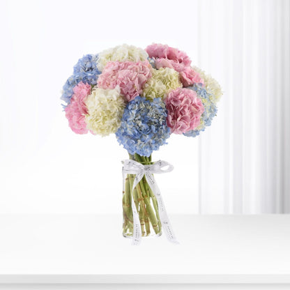 Pastel pink, blue, and cream hydrangeas in clear vase, tied with branded ribbon.