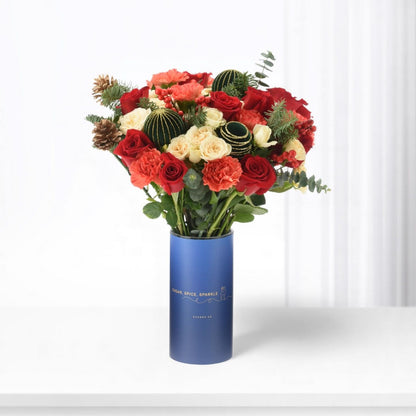 Vibrant red, cream roses, berries, pine, and ornaments in sleek blue vase.