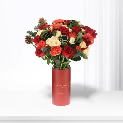 Vibrant red and cream roses, pinecones, and greenery in festive red cylinder vase.