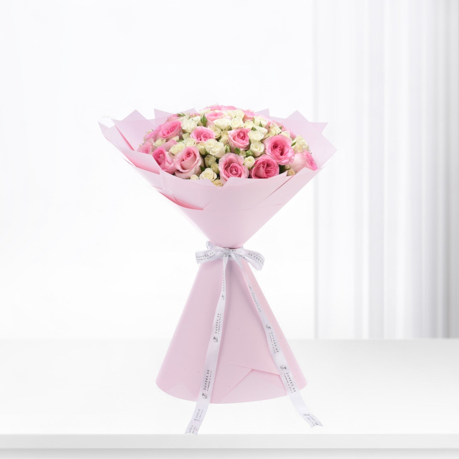 Soft pink roses and cream blooms elegantly wrapped in pastel pink paper with branded white ribbon.