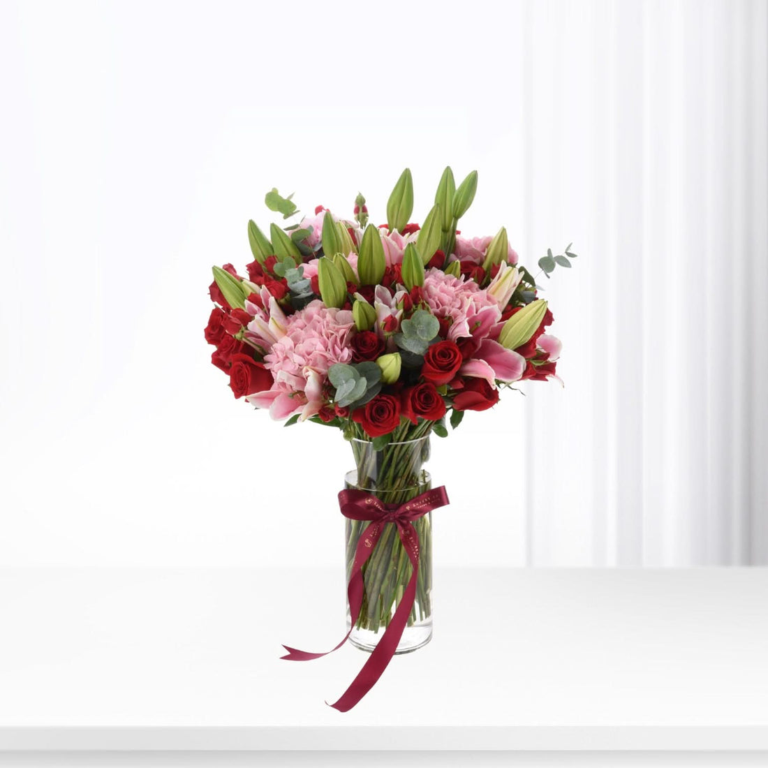 Vibrant red roses, pink hydrangeas, and green lilies in clear vase tied with burgundy ribbon.