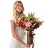 Woman holds vibrant red roses, pink hydrangeas, and green lilies in clear vase with burgundy ribbon.