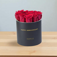 Luxury red roses in matte black "Happy Anniversary" gift box, gold lettering, elegant wooden surface.