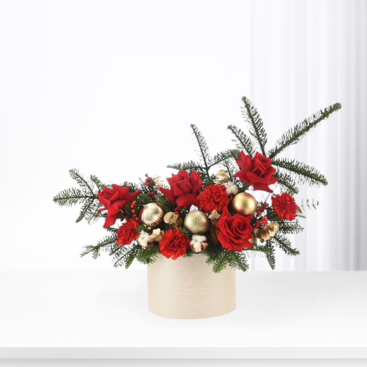 Red roses, carnations, and pine in cream box with gold ornaments.