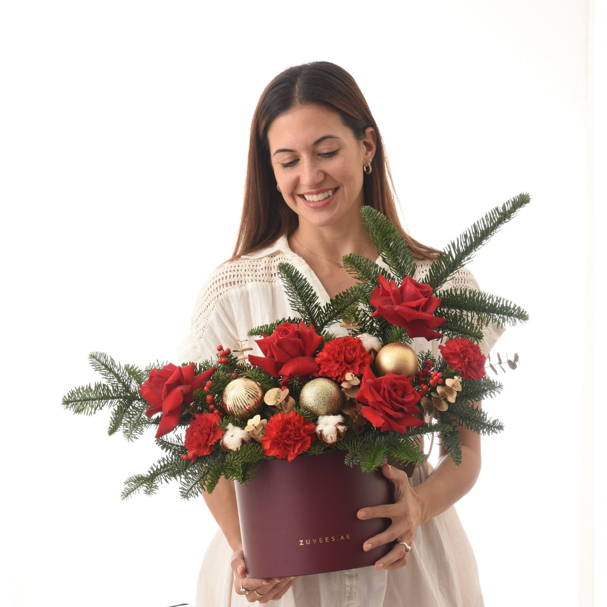 Woman holds festive red rose, carnation, pine, gold ornament bouquet in burgundy Zuvees box.