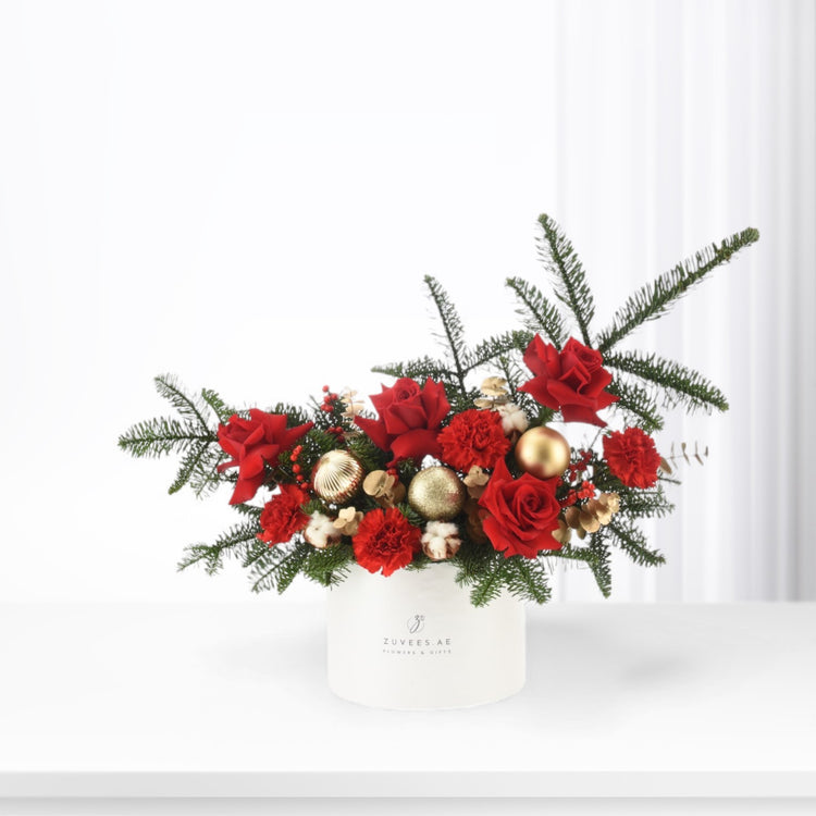 Vibrant red roses and carnations nestled in lush greenery, accented with gold ornaments, presented in a sleek white cylindrical vase.