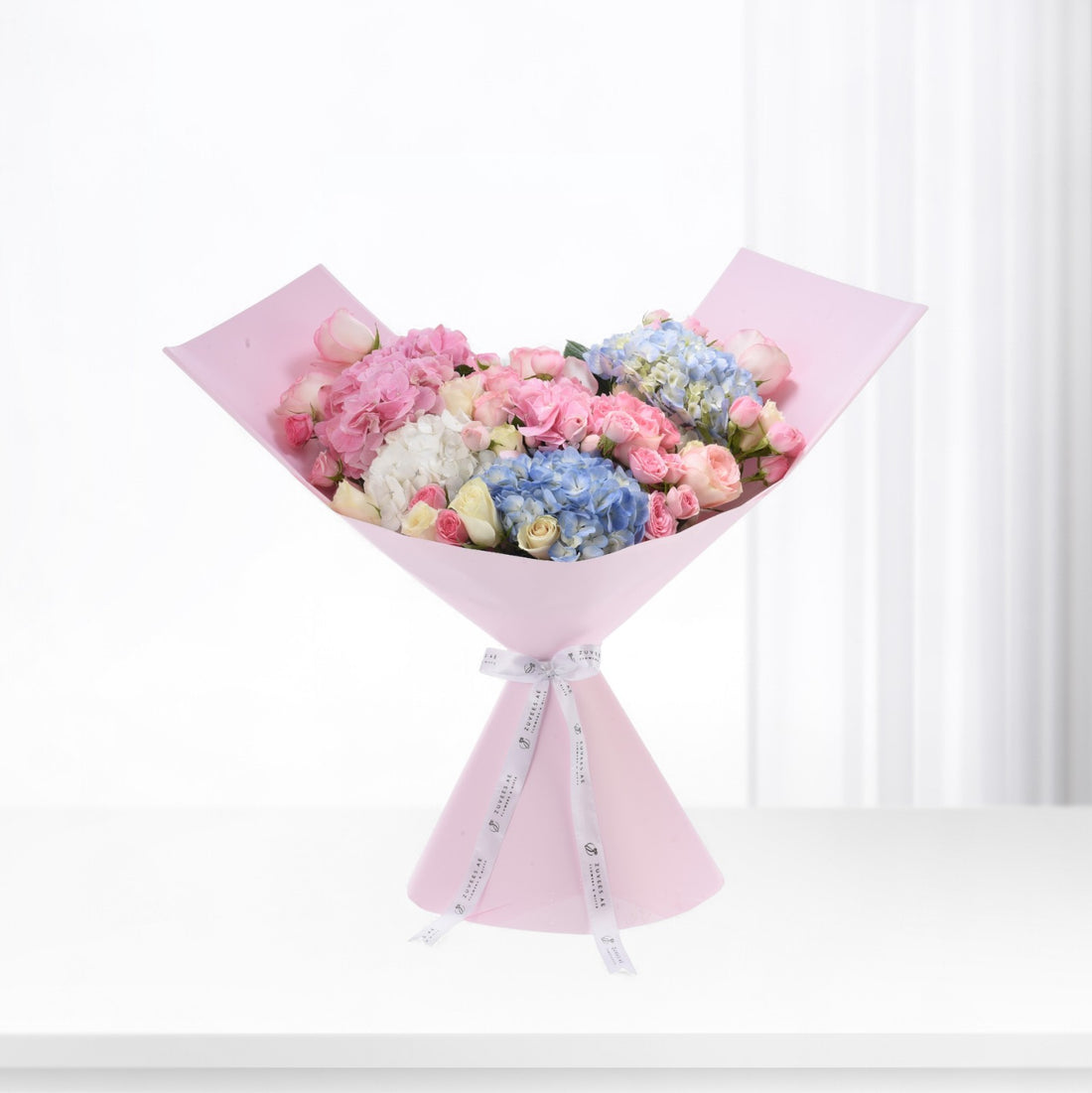 Pastel pink, blue, and white hydrangeas and roses elegantly wrapped in soft pink paper with branded ribbon.
