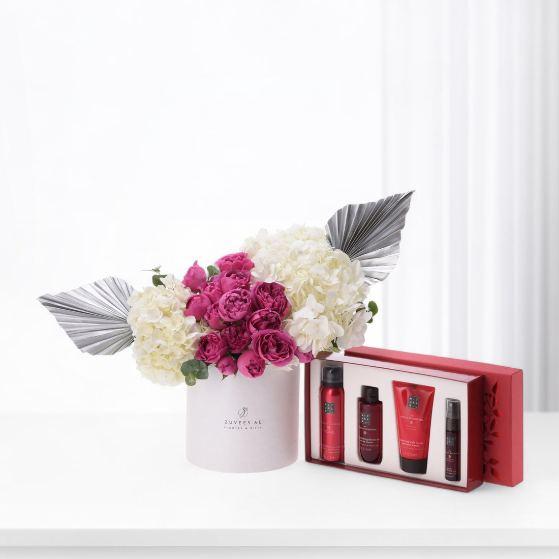 Pink roses and white hydrangeas in white vase beside red luxury skincare set.