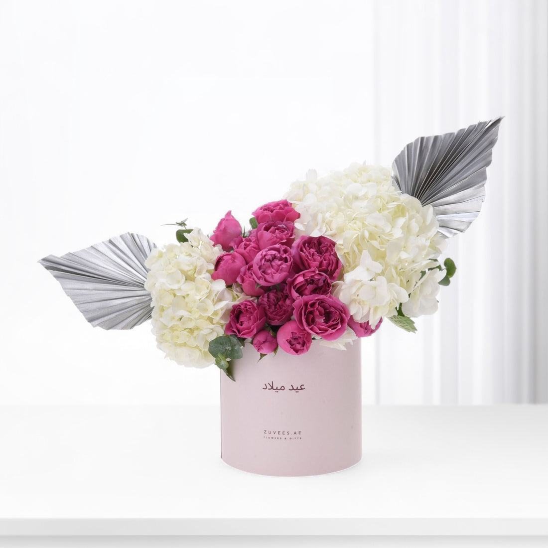 Pink box holds vibrant magenta roses and creamy hydrangeas, accented by silver fan leaves.