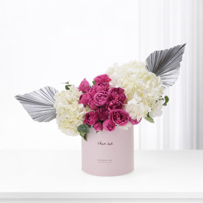 Pink box holds vibrant magenta roses and creamy hydrangeas, accented by silver fan leaves.