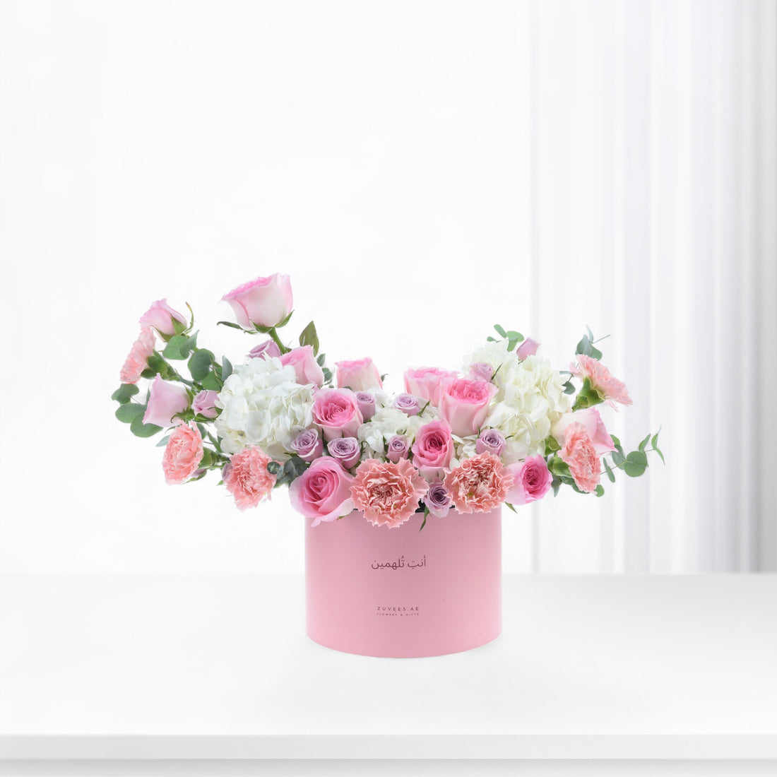 Pink roses, carnations, white hydrangeas in soft pink box with Arabic text, minimalist white background.