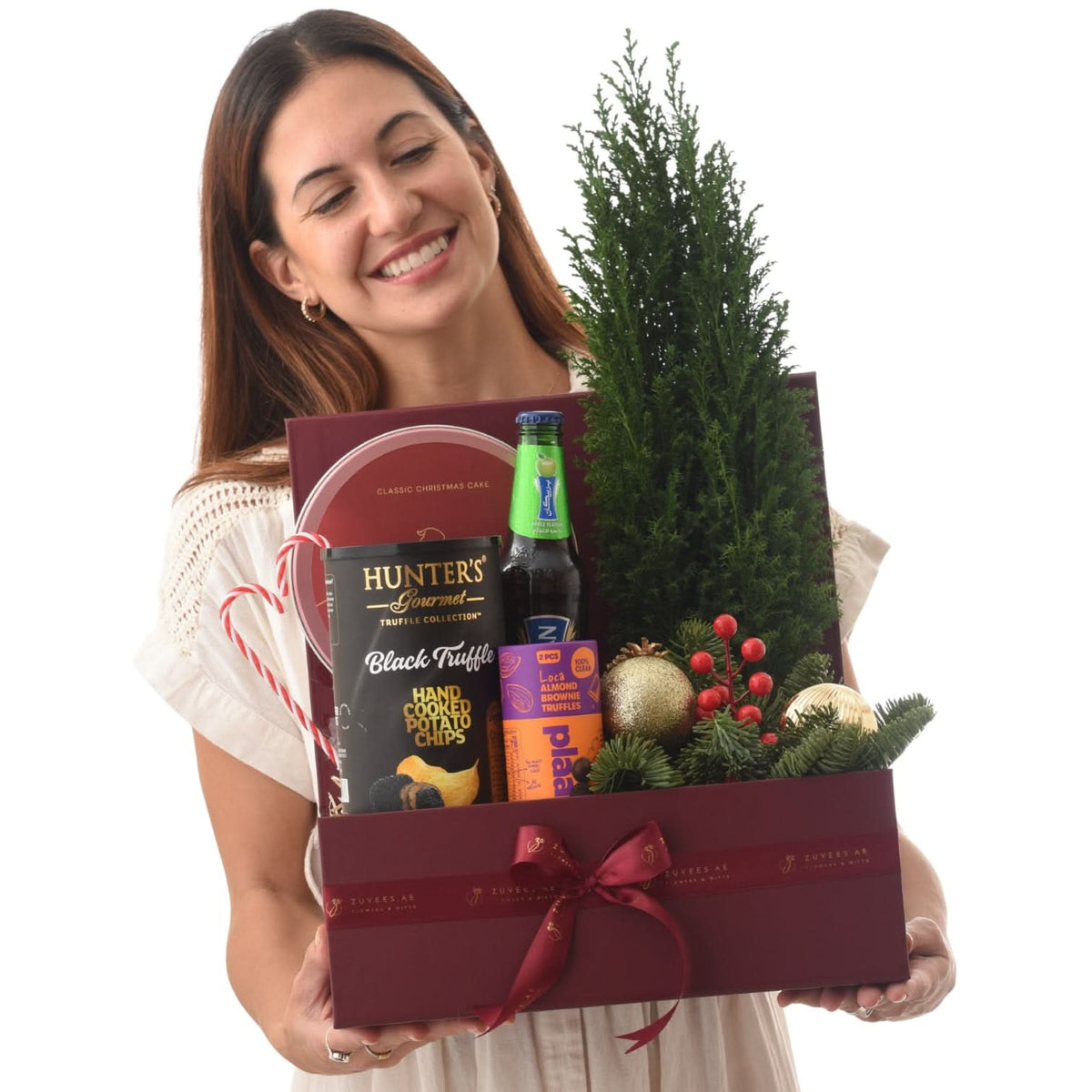 Smiling woman holds festive burgundy gift box with mini tree, truffles, cake, and drink.