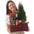 Smiling woman holds festive burgundy gift box with mini tree, truffles, cake, and drink.
