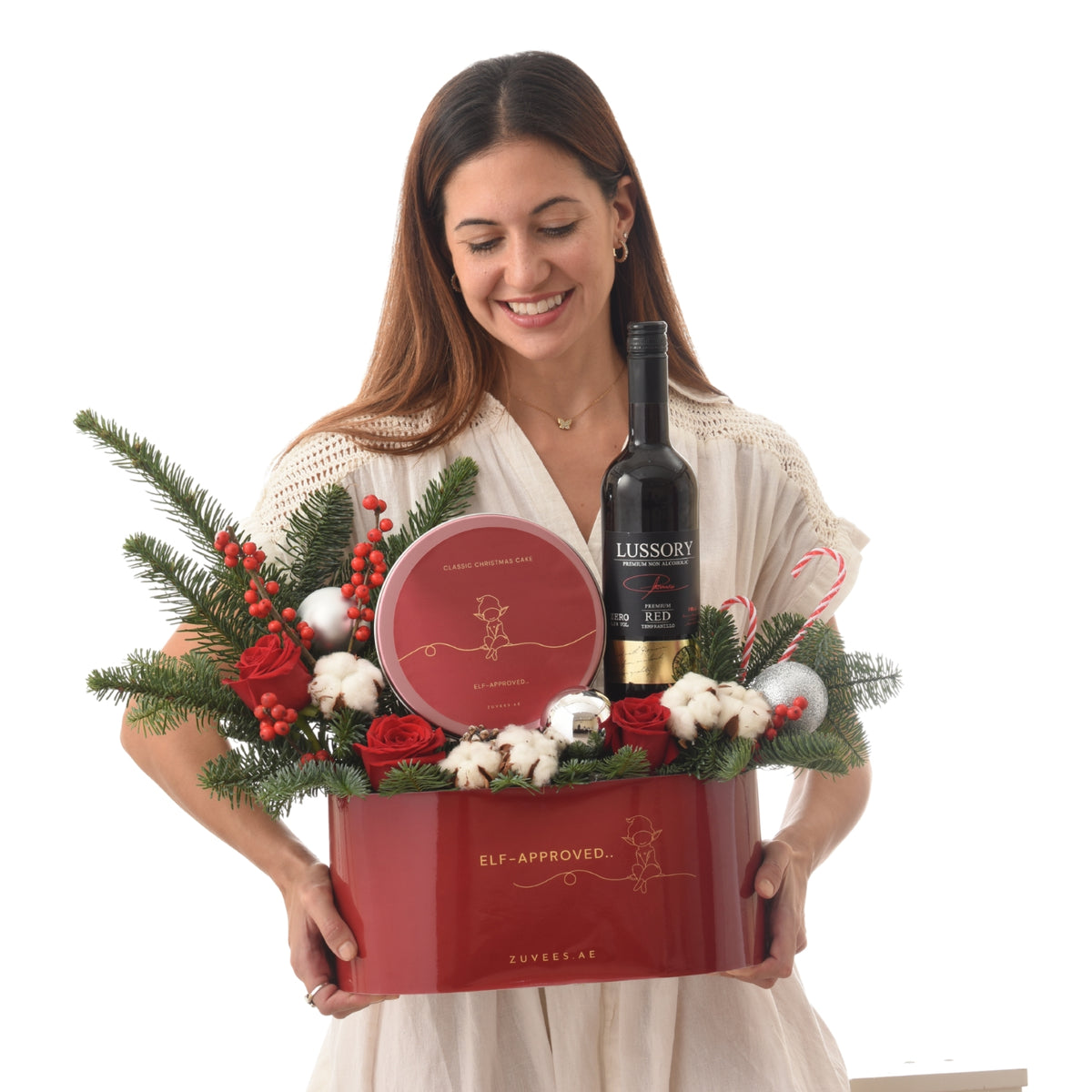 Woman holds festive red gift basket with wine, cake, roses, and holiday decor.