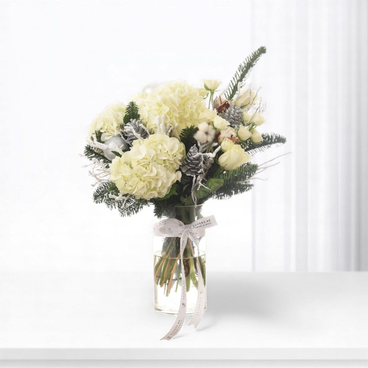 Elegant white hydrangeas, roses, and frosted pine in clear vase with branded ribbon.
