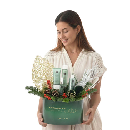Woman holds festive green gift box with pine, berries, and skincare products.