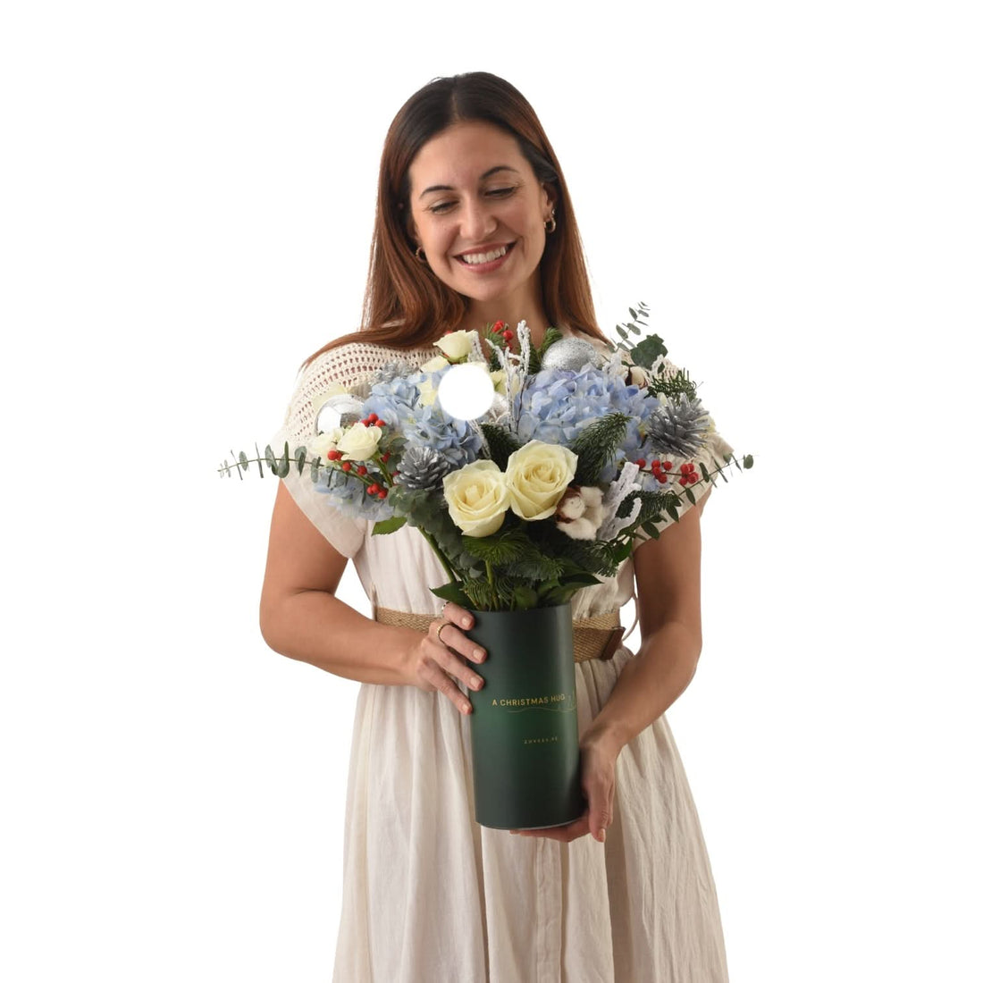 Woman smiles holding festive blue hydrangea, white rose, and silver ornament bouquet in dark green "A Christmas Hug" vase.